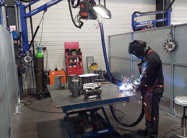 Photo d'un atelier avec un soudeur et une potence tenant en hauteur le post à souder